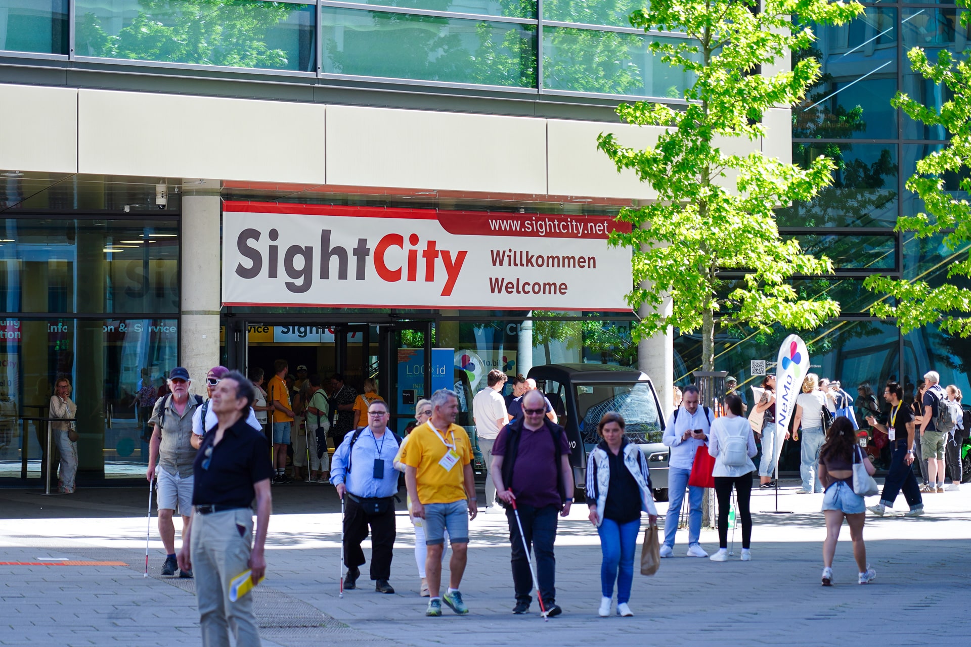 Eingang zur SihtCity-Messe. Über dem Eingang hängt ein grosses SightCity-Banner, davor laufen mehrere Personen, einige davon mit weissem Stock.