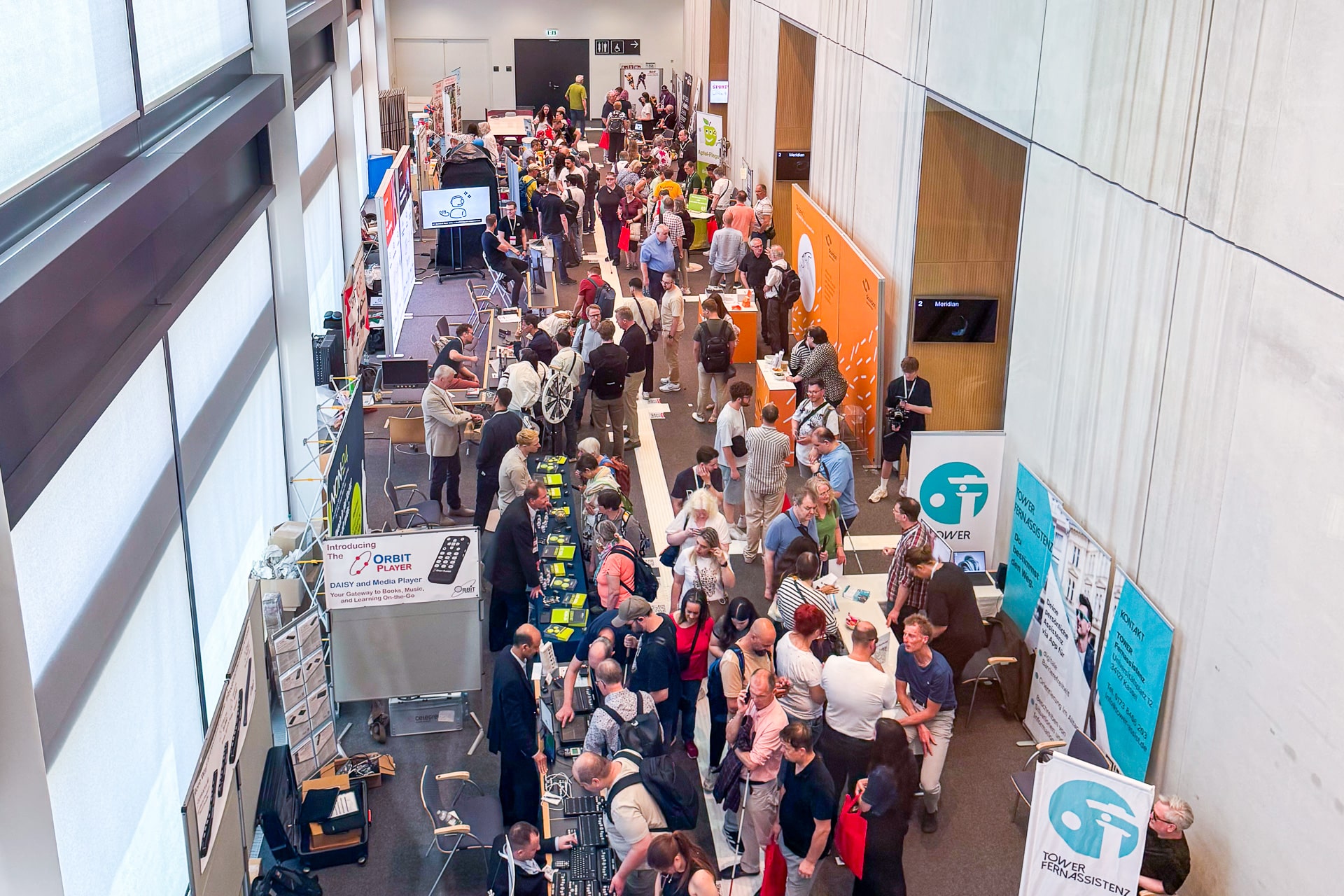 Blick von oben auf einen Teil der Messe. Links und rechts verschiedene Messestände, dazwischen laufen viele Personen.