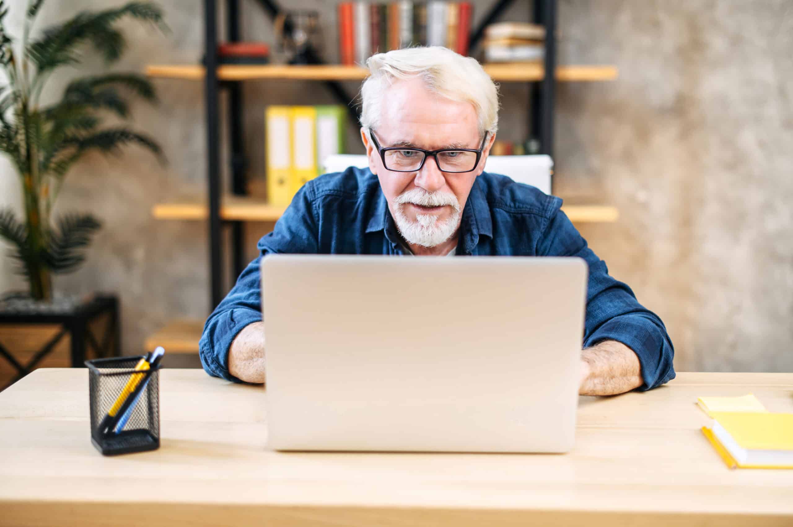 Personne âgée portant des lunettes, assise à une table, regarde attentivement, avec curiosité et un vif intérêt, un ordinateur portable ouvert. Le visage est orienté vers l’avant, le regard fixé sur l’écran. À l’arrière-plan, des étagères contenant des livres et des classeurs, dans un intérieur à lumière chaude.  