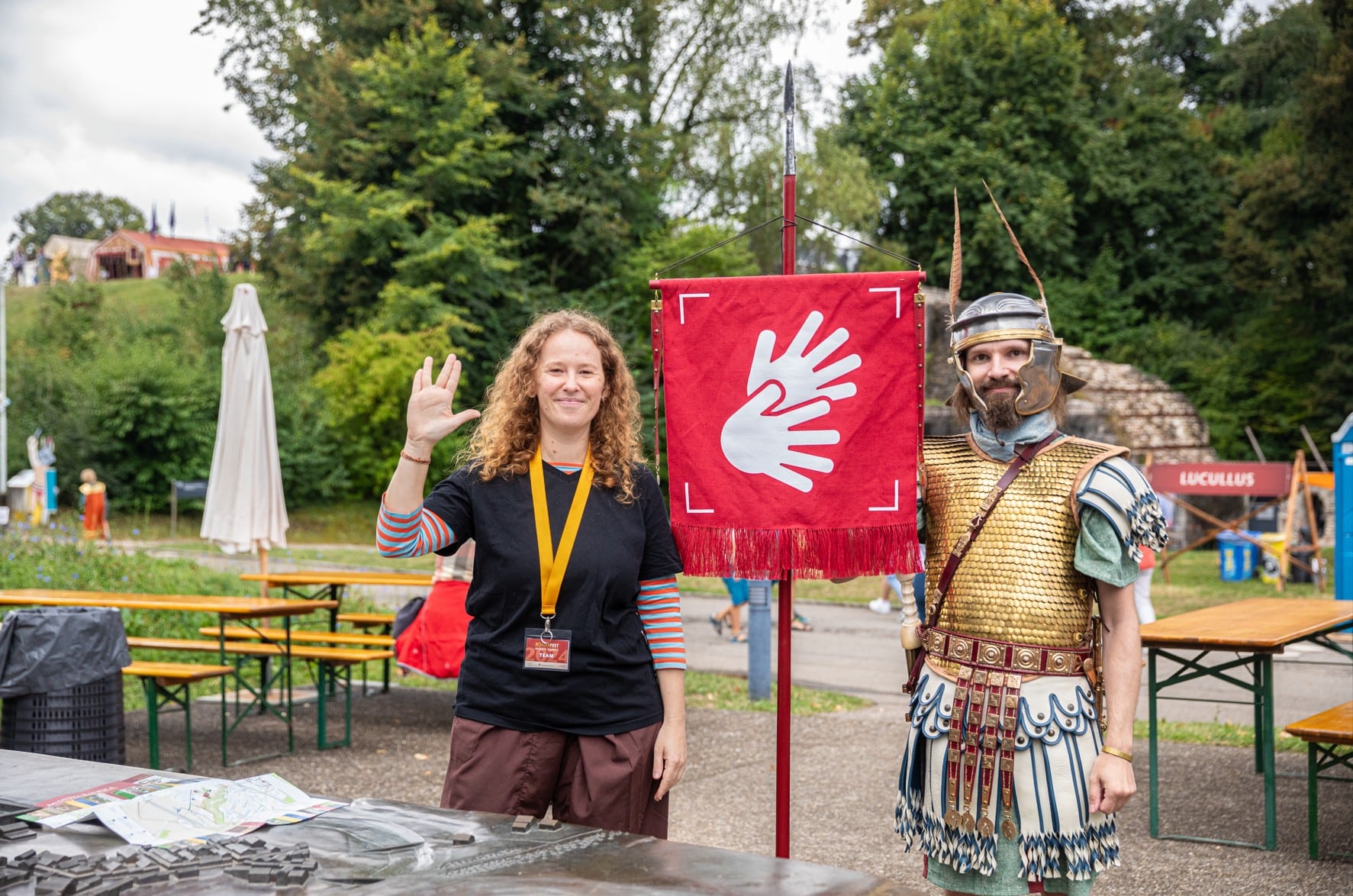 Eine Person zeigt eine Gebärde neben einem römisch gekleideten Darsteller. Vor ihnen hängt ein rotes Banner mit dem Symbol für Gebärdensprache.