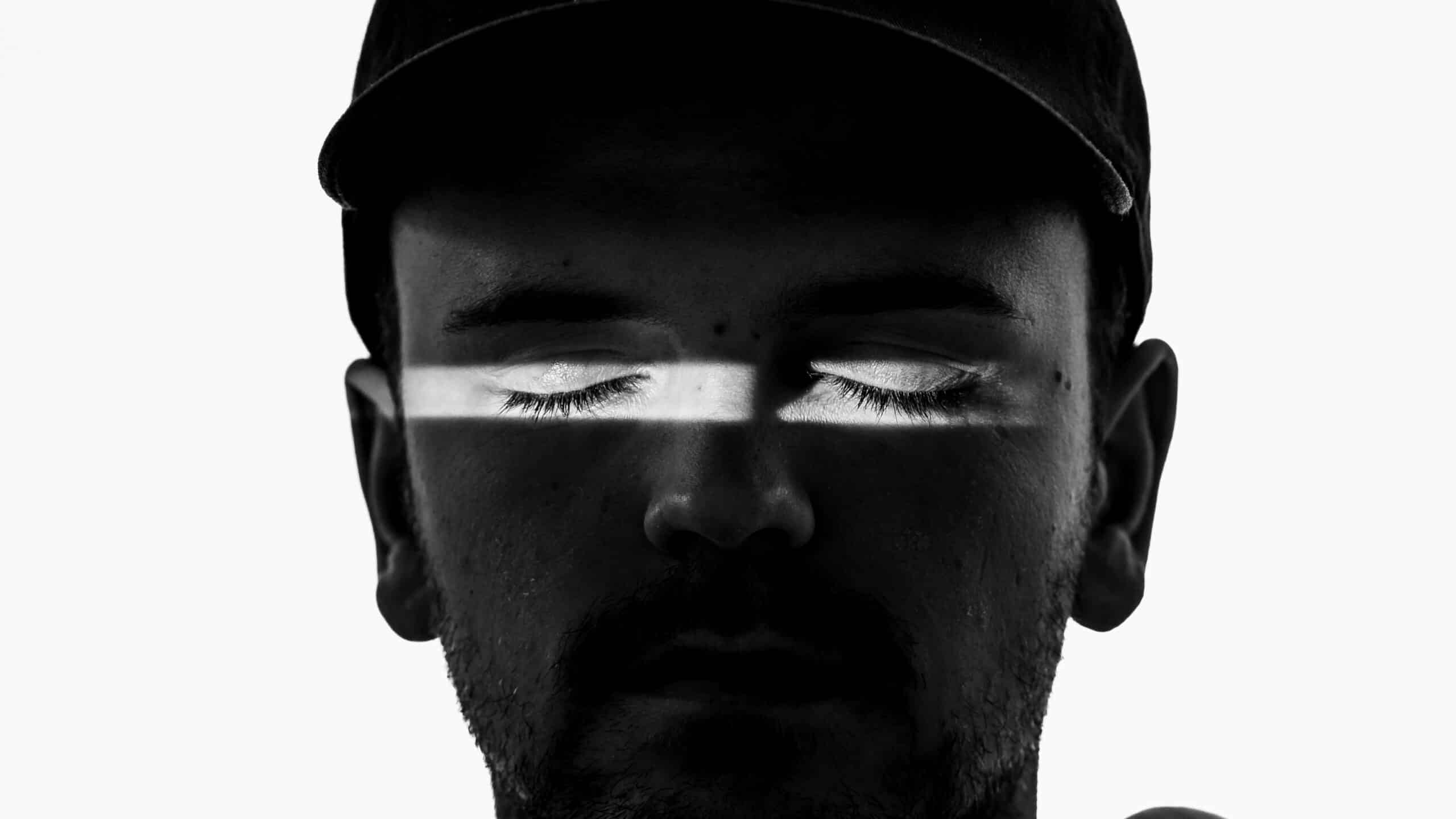 Loris Mittaz se tient face à la caméra, les yeux fermés. Il porte un chapeau foncé et un sweat à capuche clair. Seule la zone autour de ses yeux est éclairée; le reste du haut de son corps est plongé dans l'ombre.