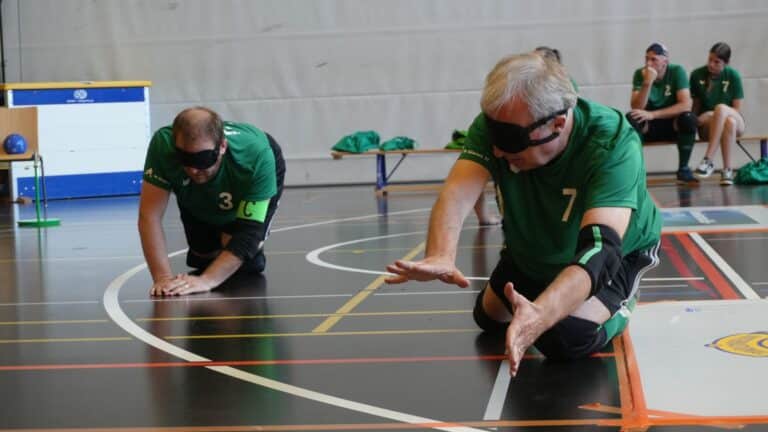 Scène tirée des STBV World Masters 2026 à Sargans : deux hommes portant des lunettes de soleil sont apparemment allongés sur le sol du gymnase après un saut ; cela semble être une scène pleine de dynamisme. À l'arrière-plan, le banc de touche où se trouvent deux autres joueurs. 