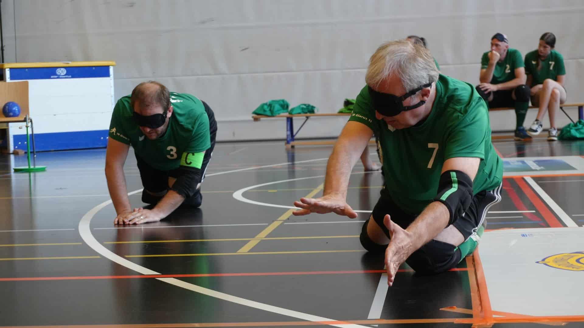 Scène tirée des STBV World Masters 2026 à Sargans : deux hommes portant des lunettes de soleil sont apparemment allongés sur le sol du gymnase après un saut ; cela semble être une scène pleine de dynamisme. À l'arrière-plan, le banc de touche où se trouvent deux autres joueurs. 
