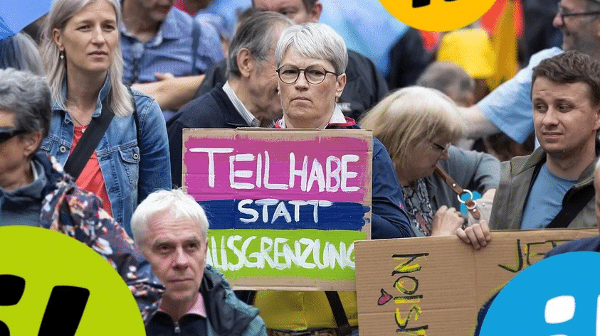 Frau hält in einer Menschenmenge ein Schild mit der Aufschrift «Teilhabe statt Ausgrenzung» hoch.