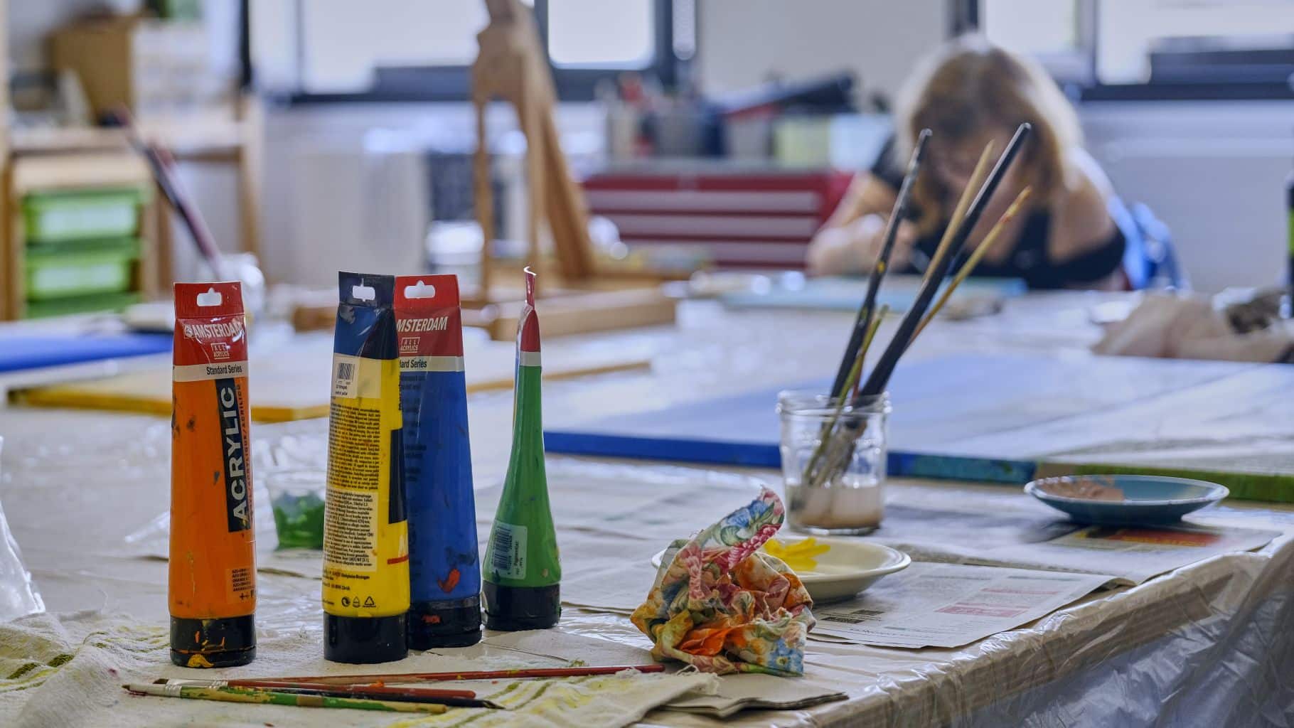 Ästhetisches Bild Pinsel und verschiedene Farben auf einem Tisch voller Farbkleckse, im Hintergruns sieht man unscharf einen grossen hellen Raum mit grossem Tisch und eine Person, die gerade malt