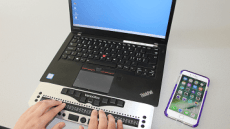 Ein Laptop mit einer Braillezeile auf der Finger etwas ertasten. Daneben ein entsperrtes iPhone.