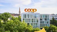 Foto des Coop-Hauptsitzes in Basel. Auf dem Dach prangt gross der Schriftzug Coop. Im Hintergrund links ist eine Kirche zu erkennen, im Vordergrund grüne Bäume. 