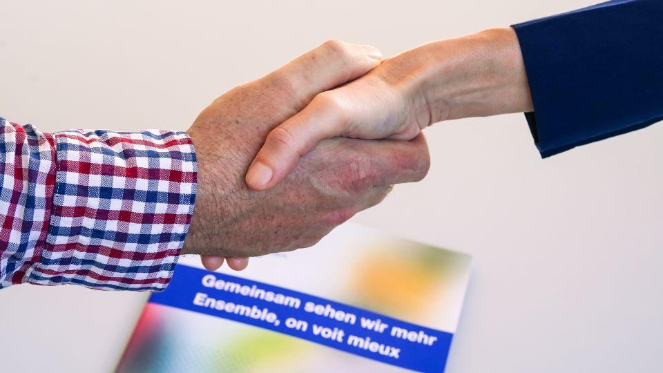 Zwei Hände machen einen Handschlag. Dahinter ist ein SBV-Flyer mit dem Slogan "Gemeinsam sehen wir mehr" zu sehen.