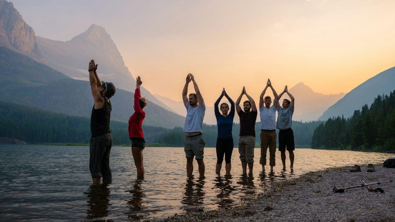 Un groupe de personnes en position de yoga, les pieds dans l’eau, avec, à l’arrière-plan, des montagnes et le soleil levant.